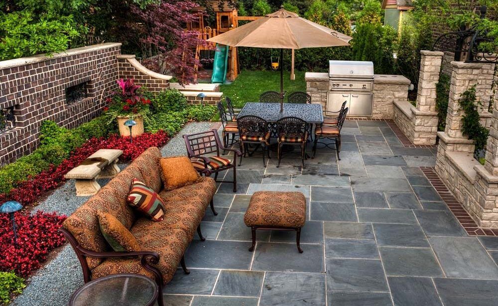 Patio with outdoor dining set, sofa, grill, and stone pavers surrounded by greenery and brick wall.