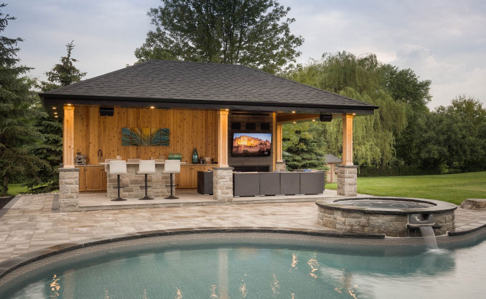 Poolside cabana with bar, TV, and hot tub. Grey stone patio, dark roof, trees in the background.