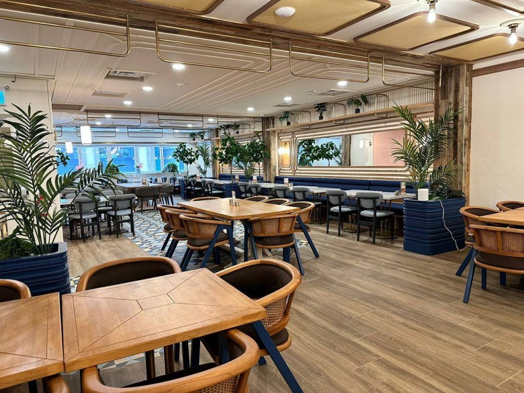 Restaurant interior with wooden tables and chairs, blue accents, plants, and a large mirror.
