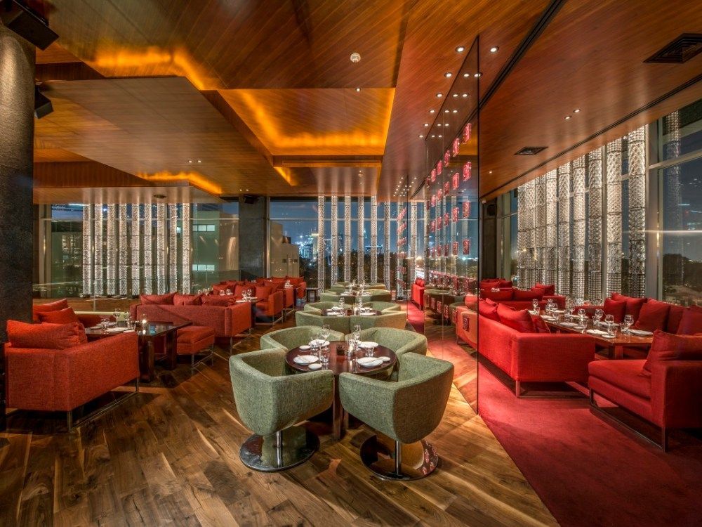 Upscale restaurant interior with red and green seating, wooden floors and ceiling, and city view at night.