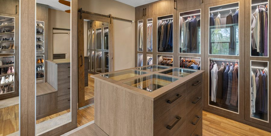 Walk-in closet with brown wood cabinets, clothing, glass display, and full-length mirror.