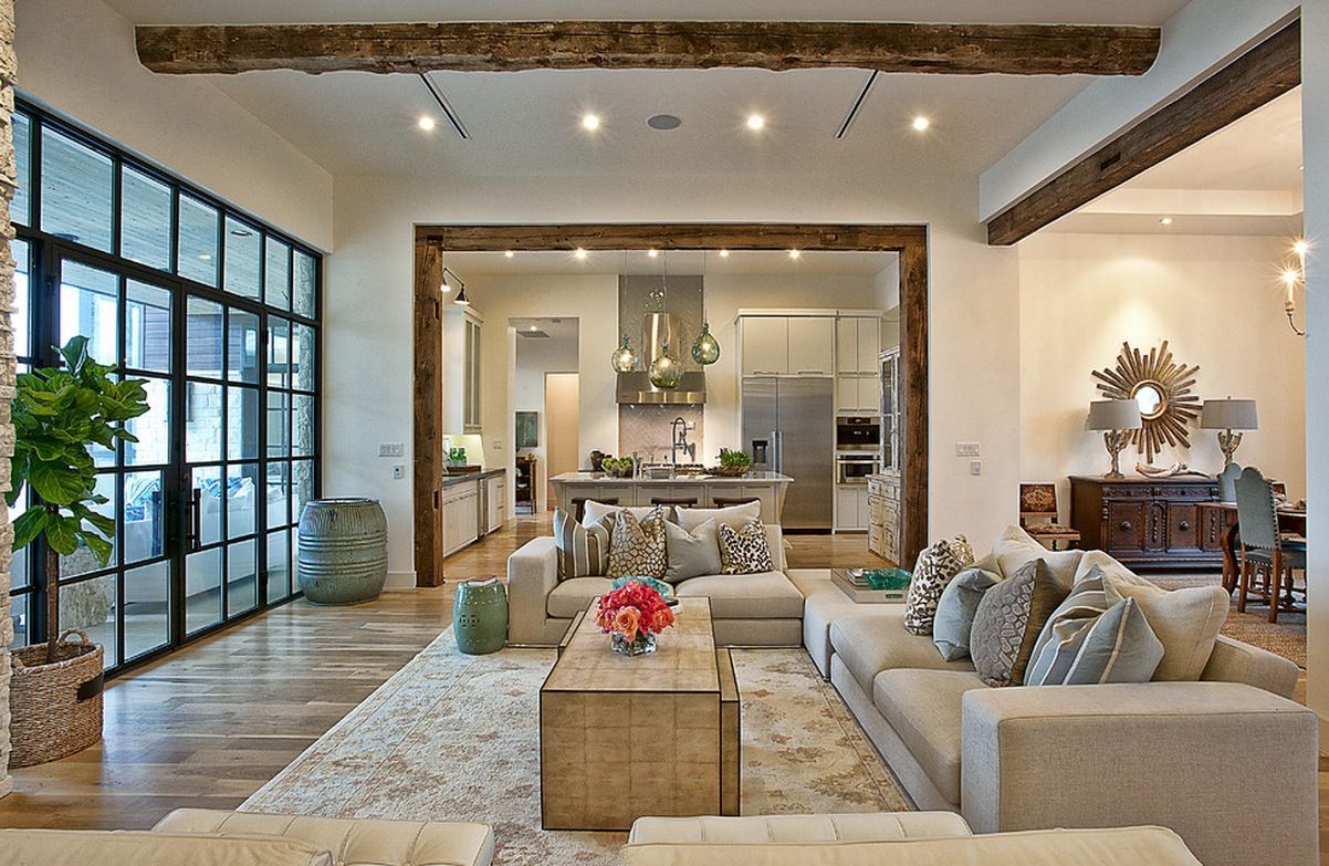 Open-concept living room with neutral-toned sofa, wooden beams, large window, and view into the kitchen.
