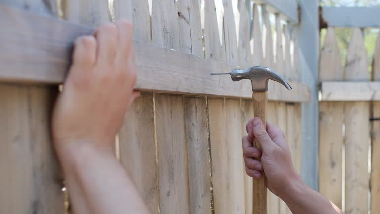 Hands hammering a nail into a wooden fence, securing a horizontal board.
