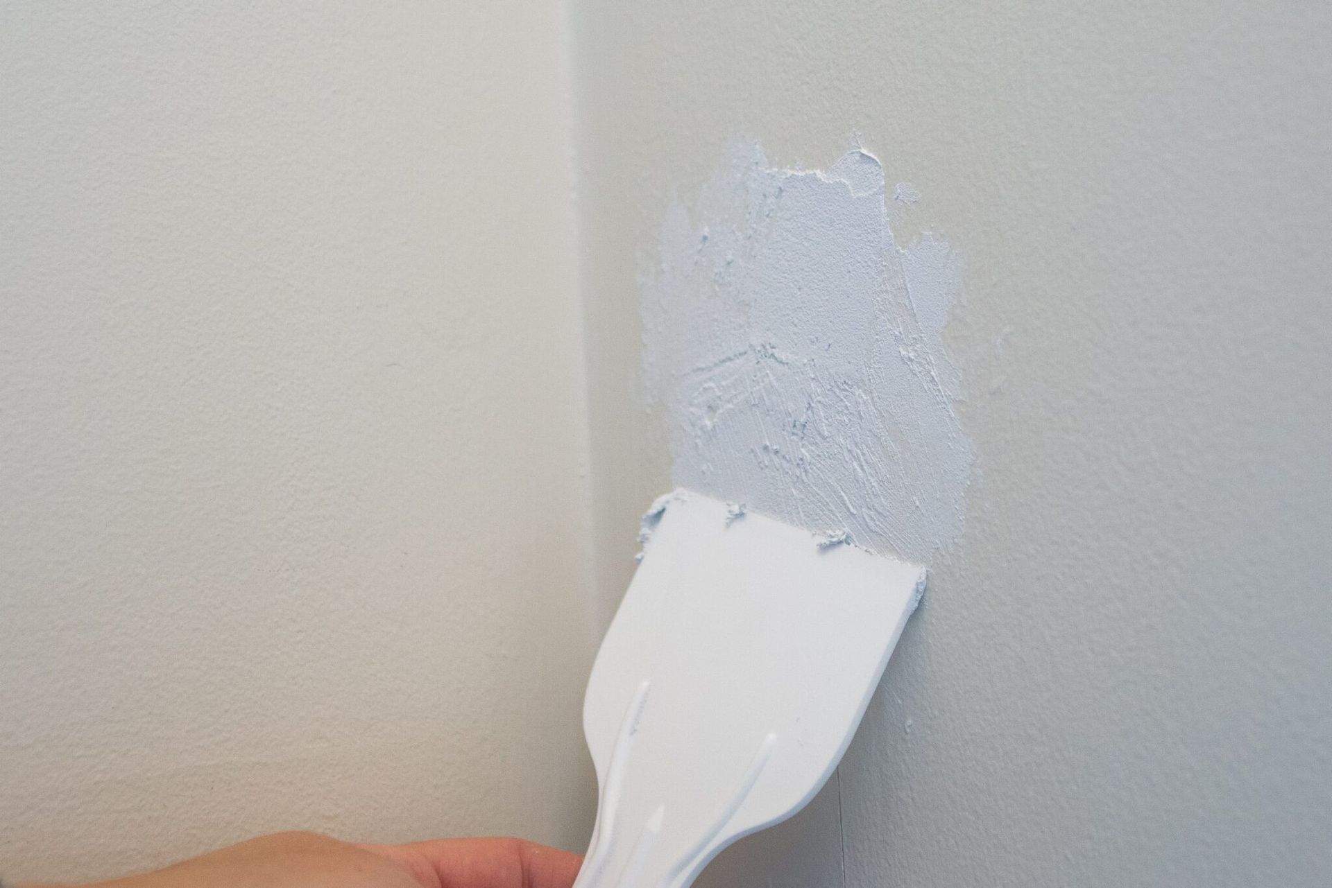 A person uses a putty knife to apply light blue spackle to a wall corner.