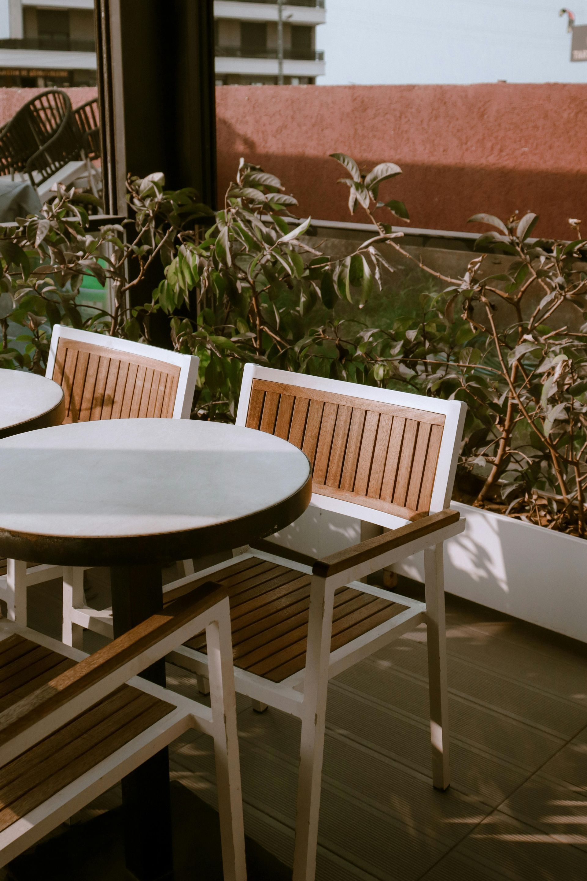 Outdoor cafe seating with wooden chairs and round tables, surrounded by plants.