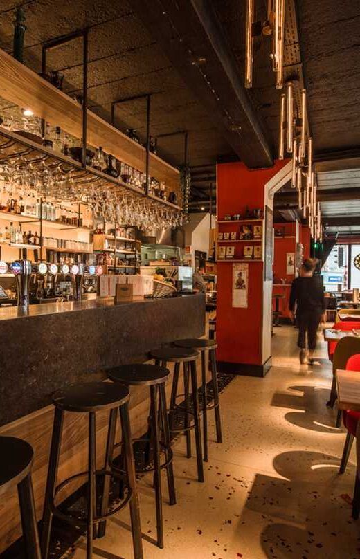 Restaurant interior with bar stools, bar, shelves with glasses and bottles, person walking.