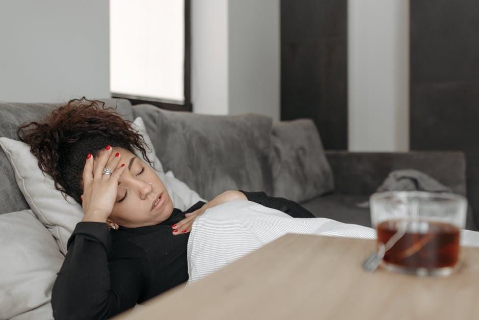 Woman with curly hair rests on a sofa, looking unwell; a cup of tea on a table beside her.