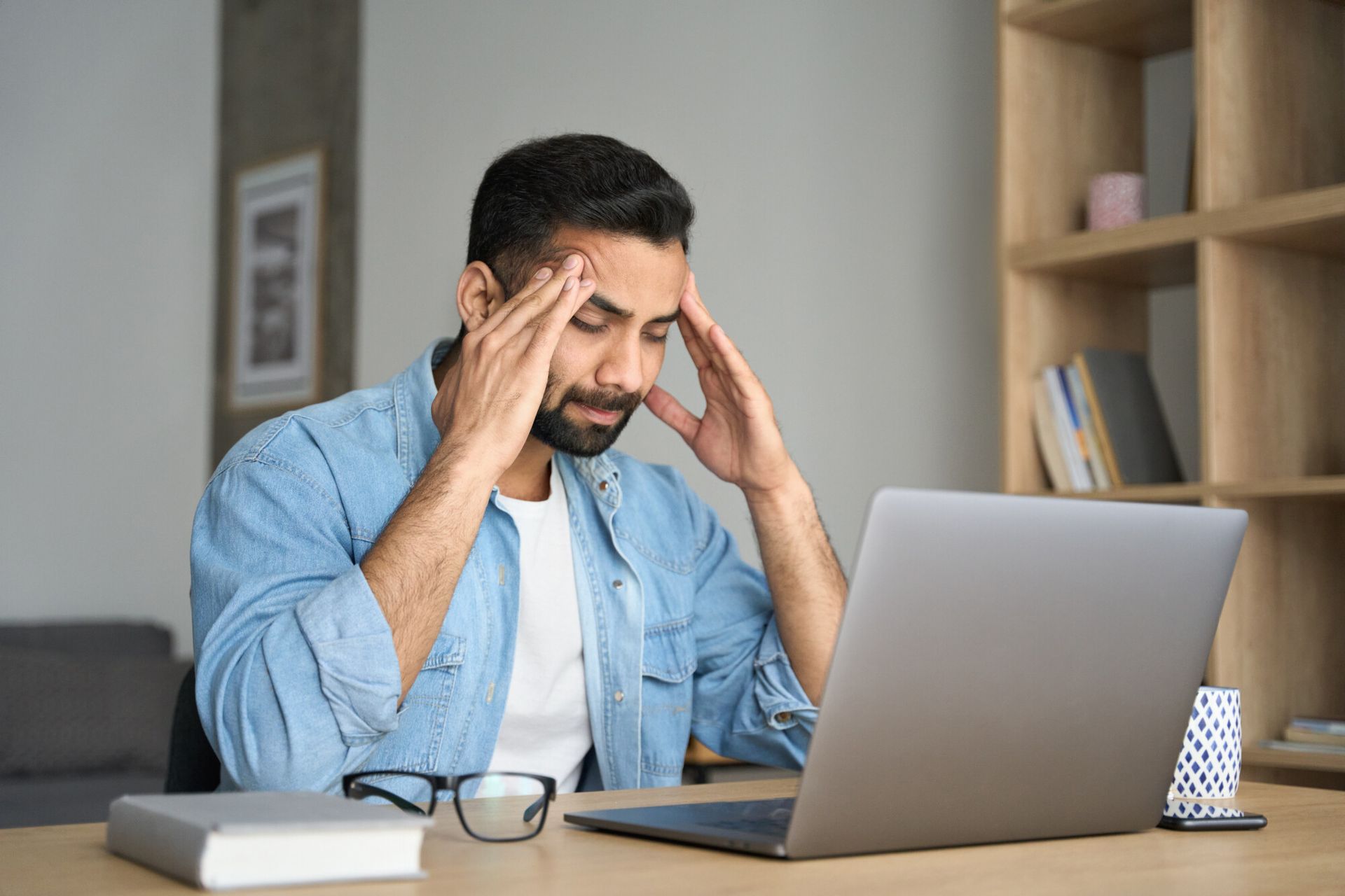 Man with a beard, holding his head, looking stressed, sitting at a laptop.