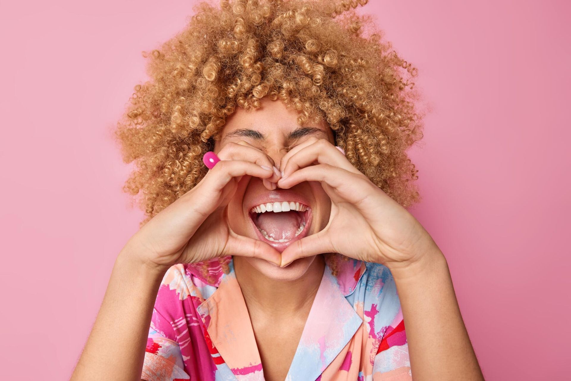 Woman with curly blonde hair makes heart shape with hands, laughing, against a pink background.