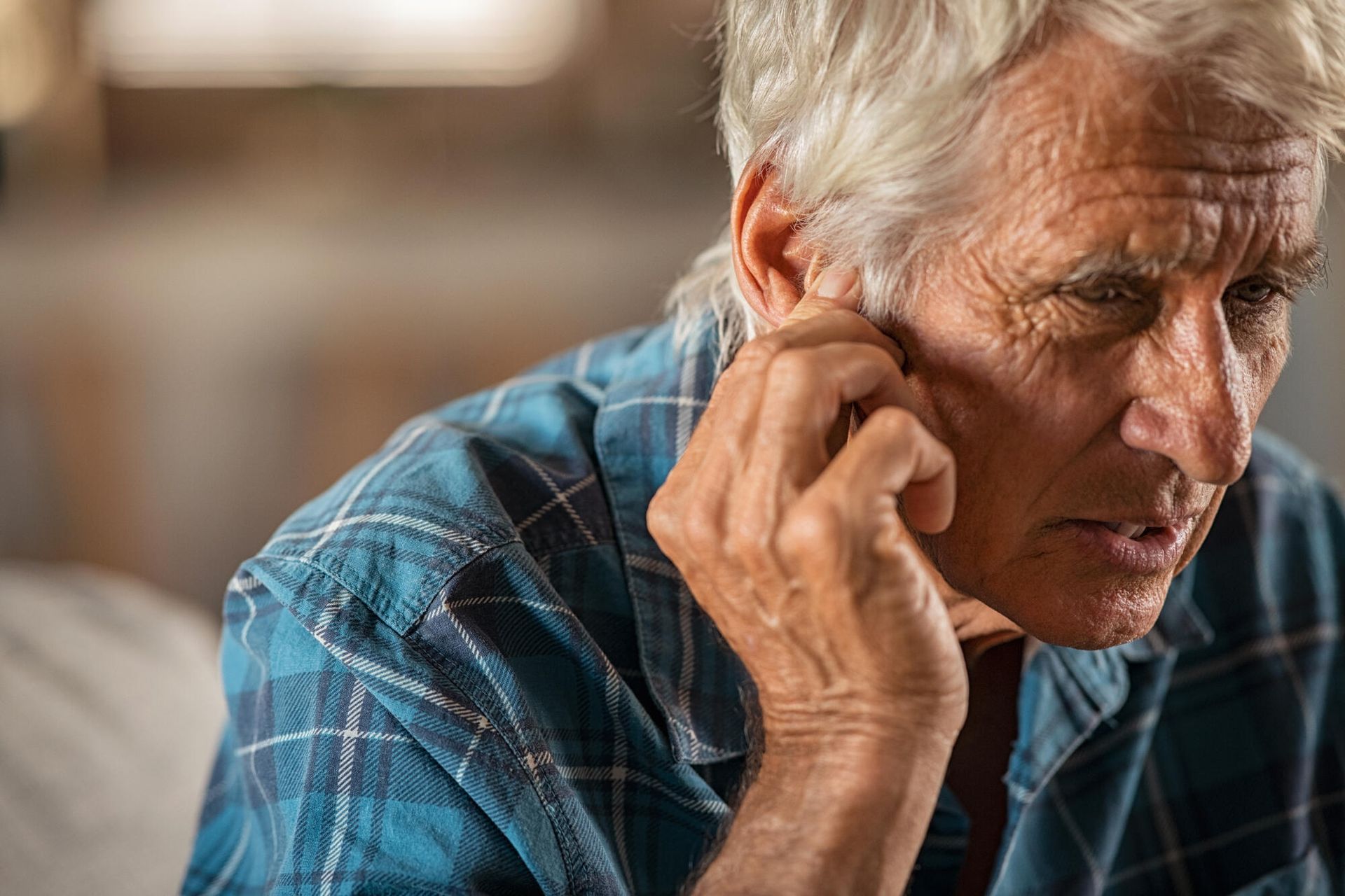 Older person with hand near ear, possibly listening. Wearing plaid shirt.