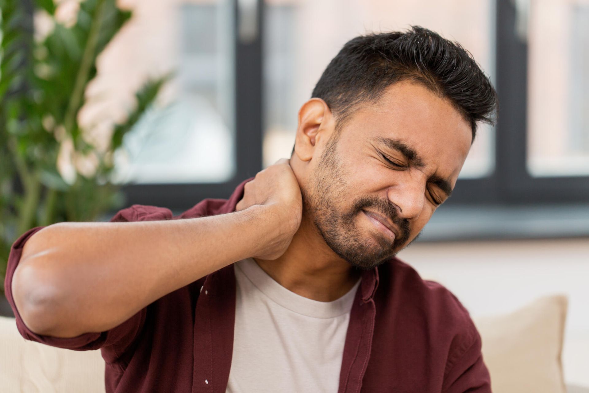 A man is sitting on a couch holding his neck in pain.