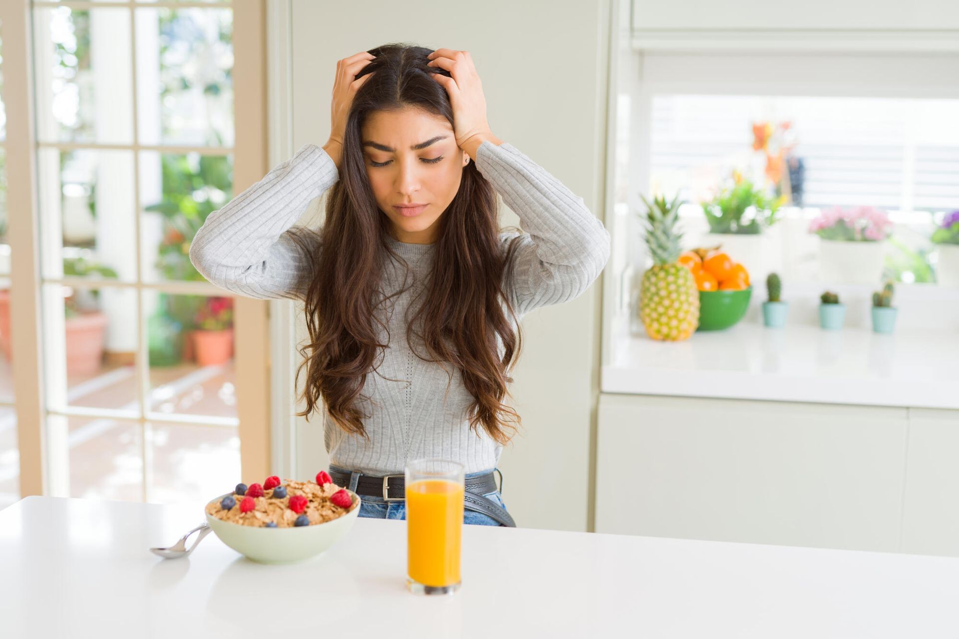 Woman holding head, looking down, next to cereal, juice, and fruit.