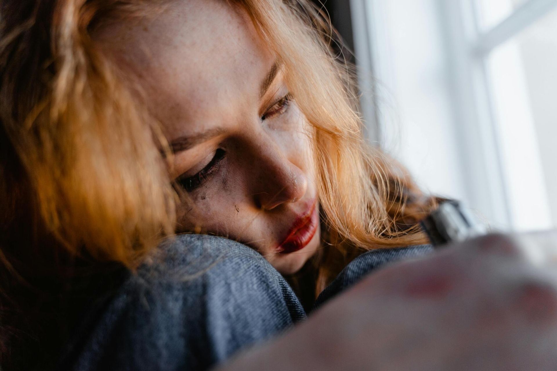 Woman with red hair, leaning against a window, with tear-streaked makeup and a somber expression.