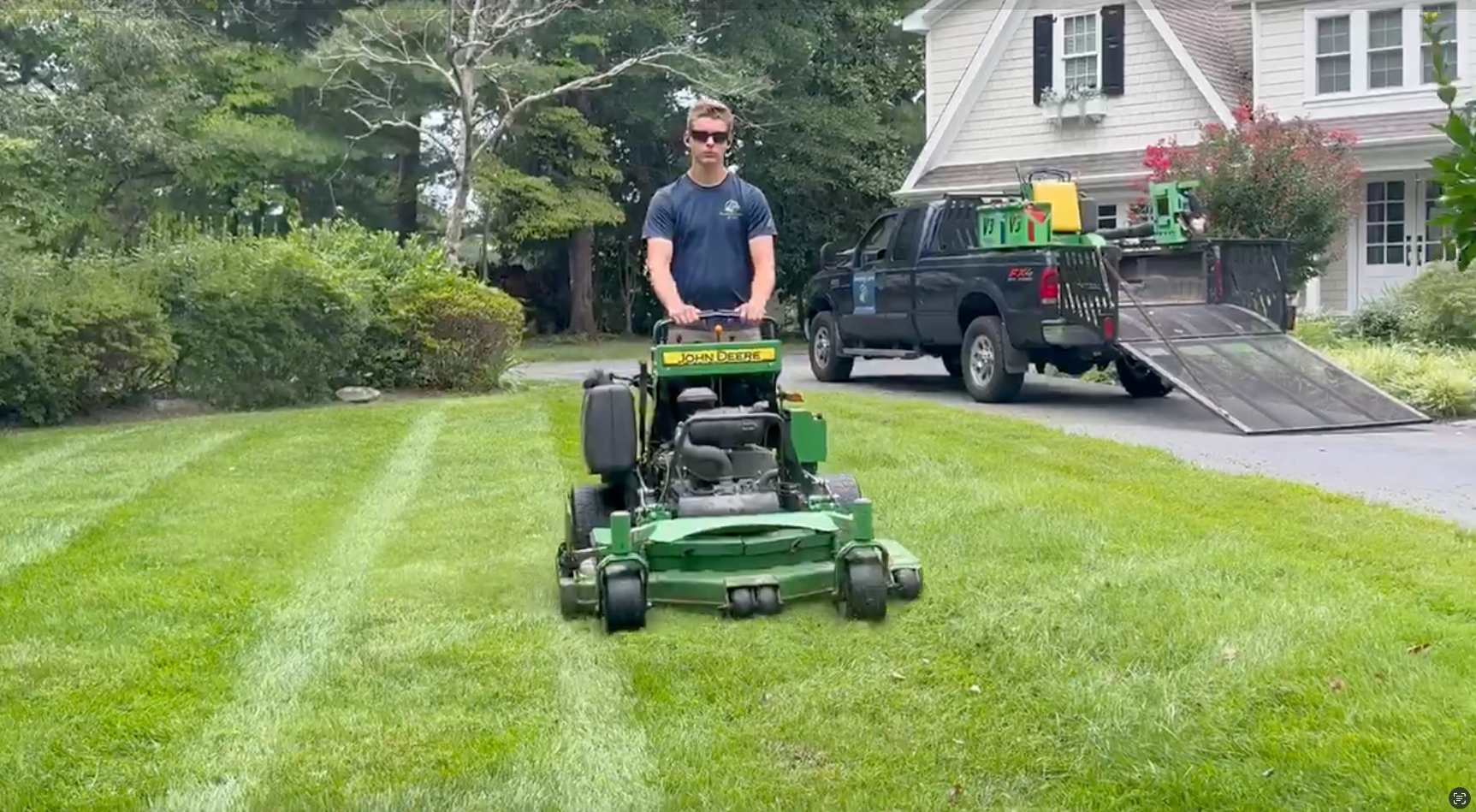 Man mowing a striped green lawn on a riding mower in front of a house with a trailer ramp nearby