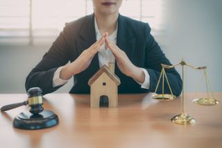 A woman is sitting at a table with a wooden house , scales of justice , and a gavel.