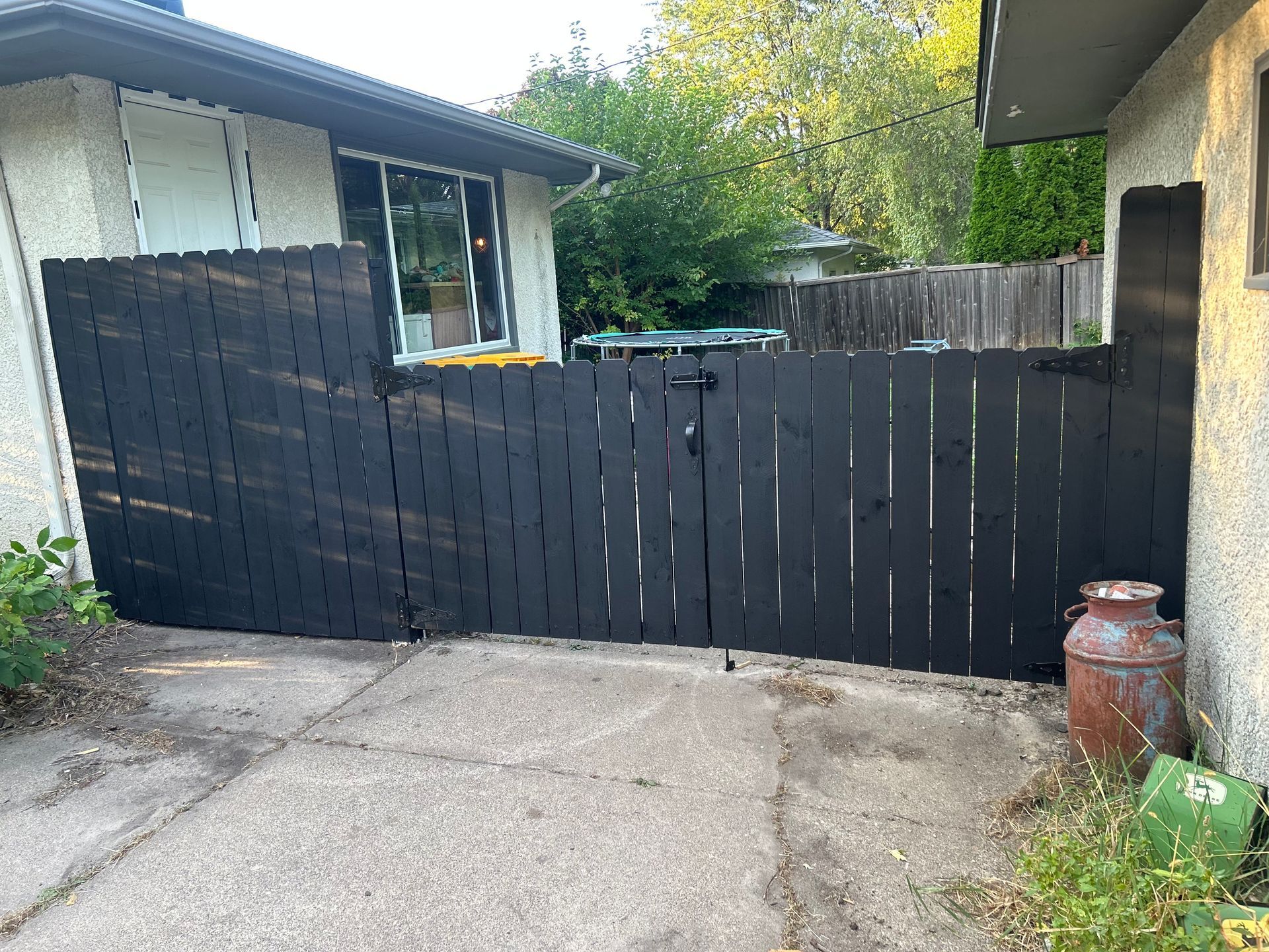 Black wooden fence in front of a house, blocking off a driveway. An old can sits to the right.
