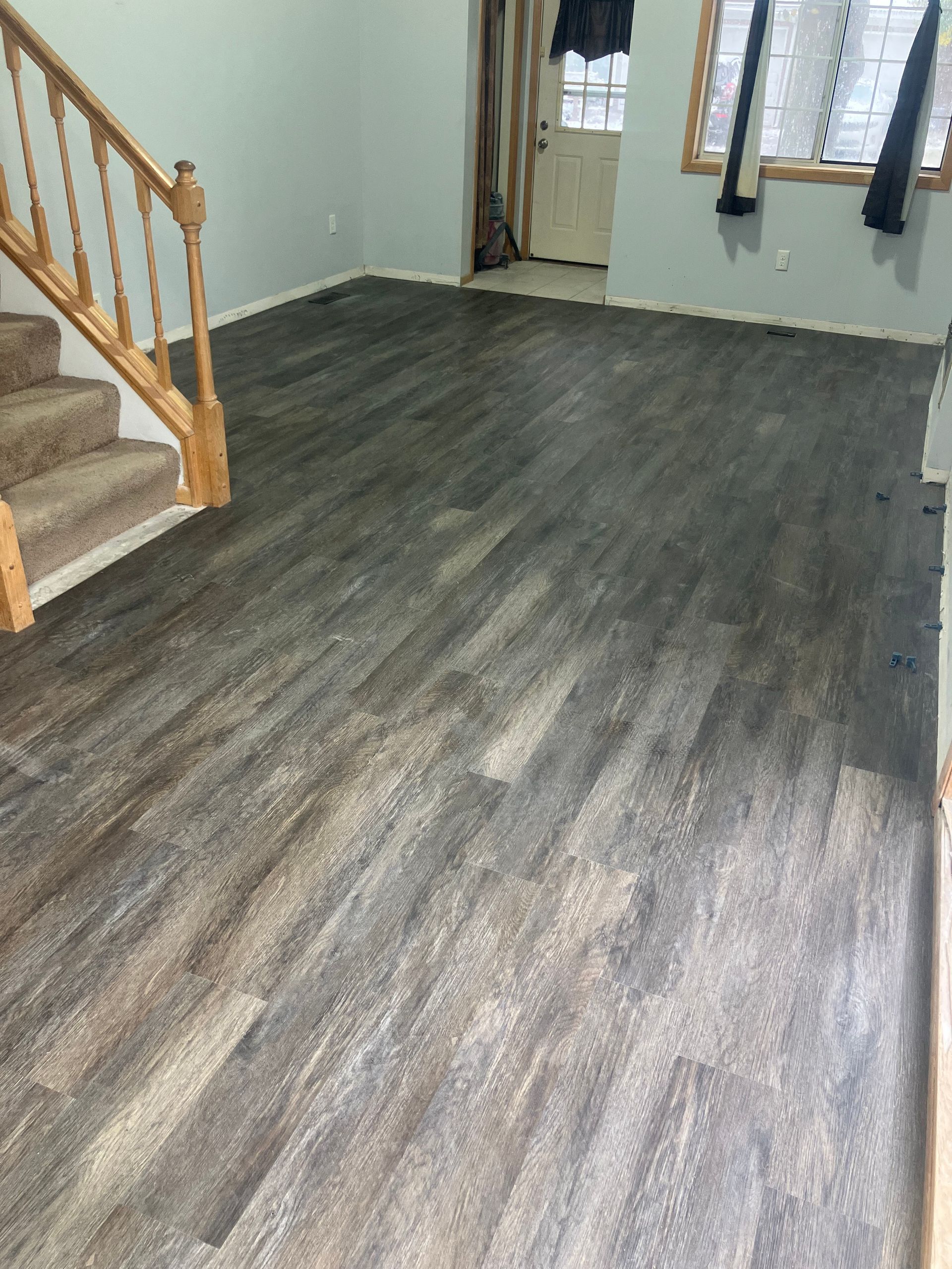 A room with newly installed dark wood-look flooring. Staircase on the left. Light walls, door and window in the background.