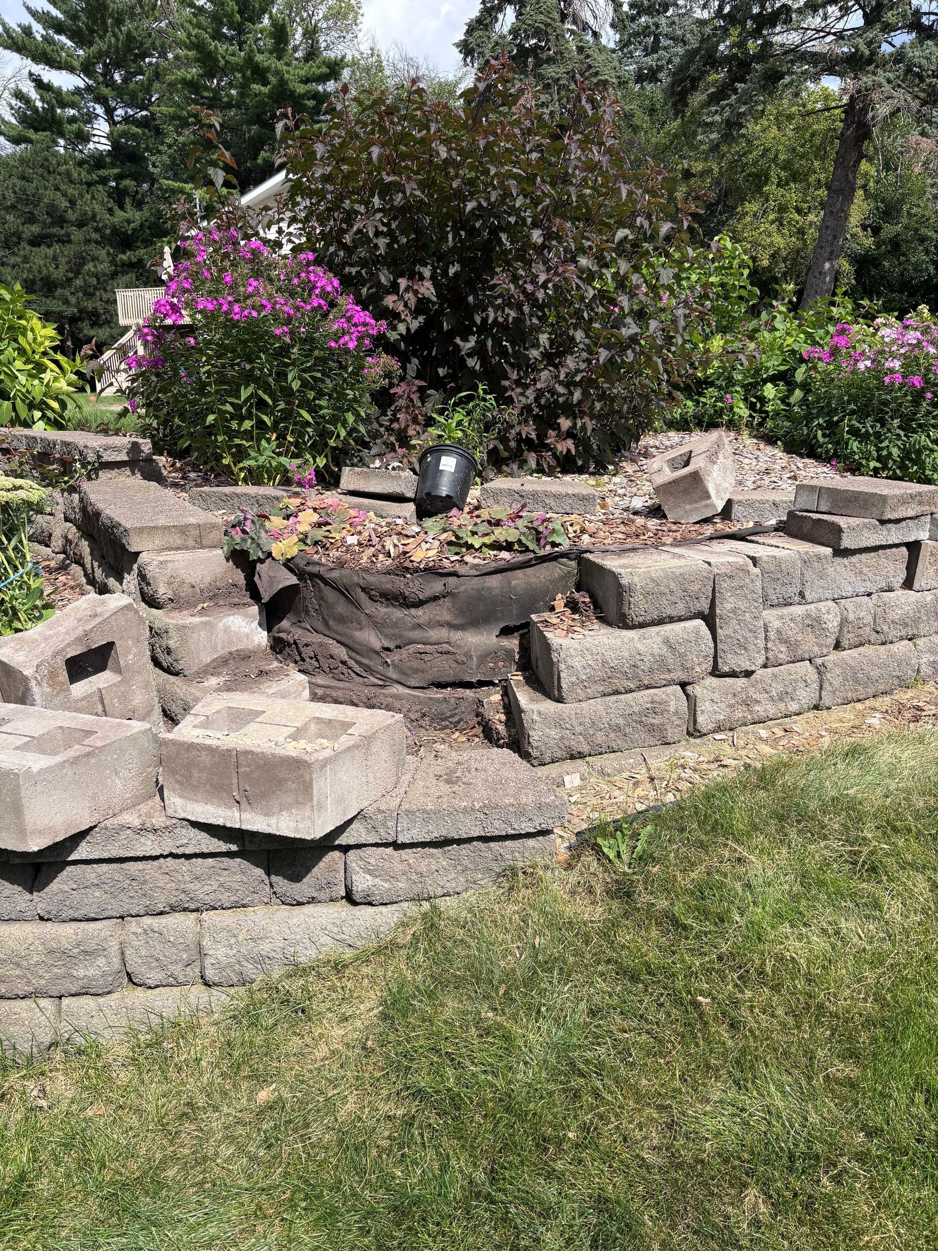 Damaged retaining wall made of gray blocks in a garden with colorful flowers.
