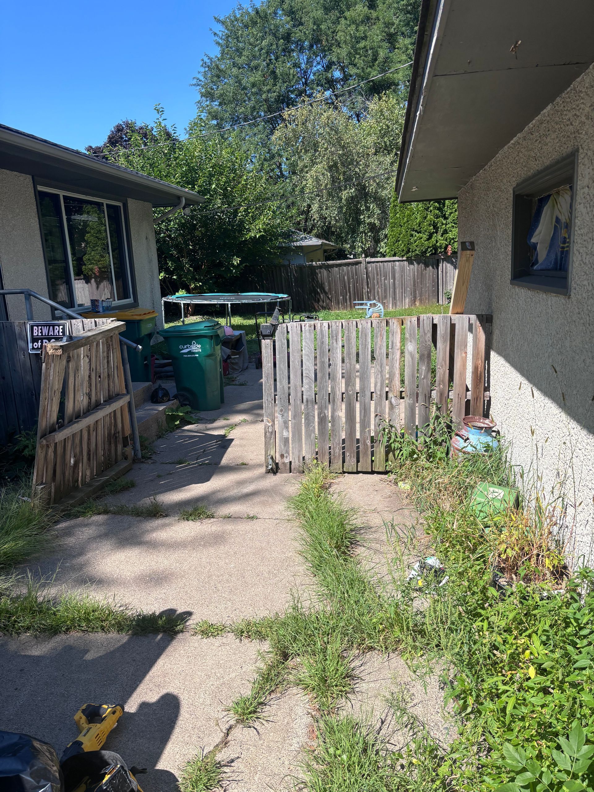 A concrete path overgrown with weeds between two buildings, a fence and trash in the back.
