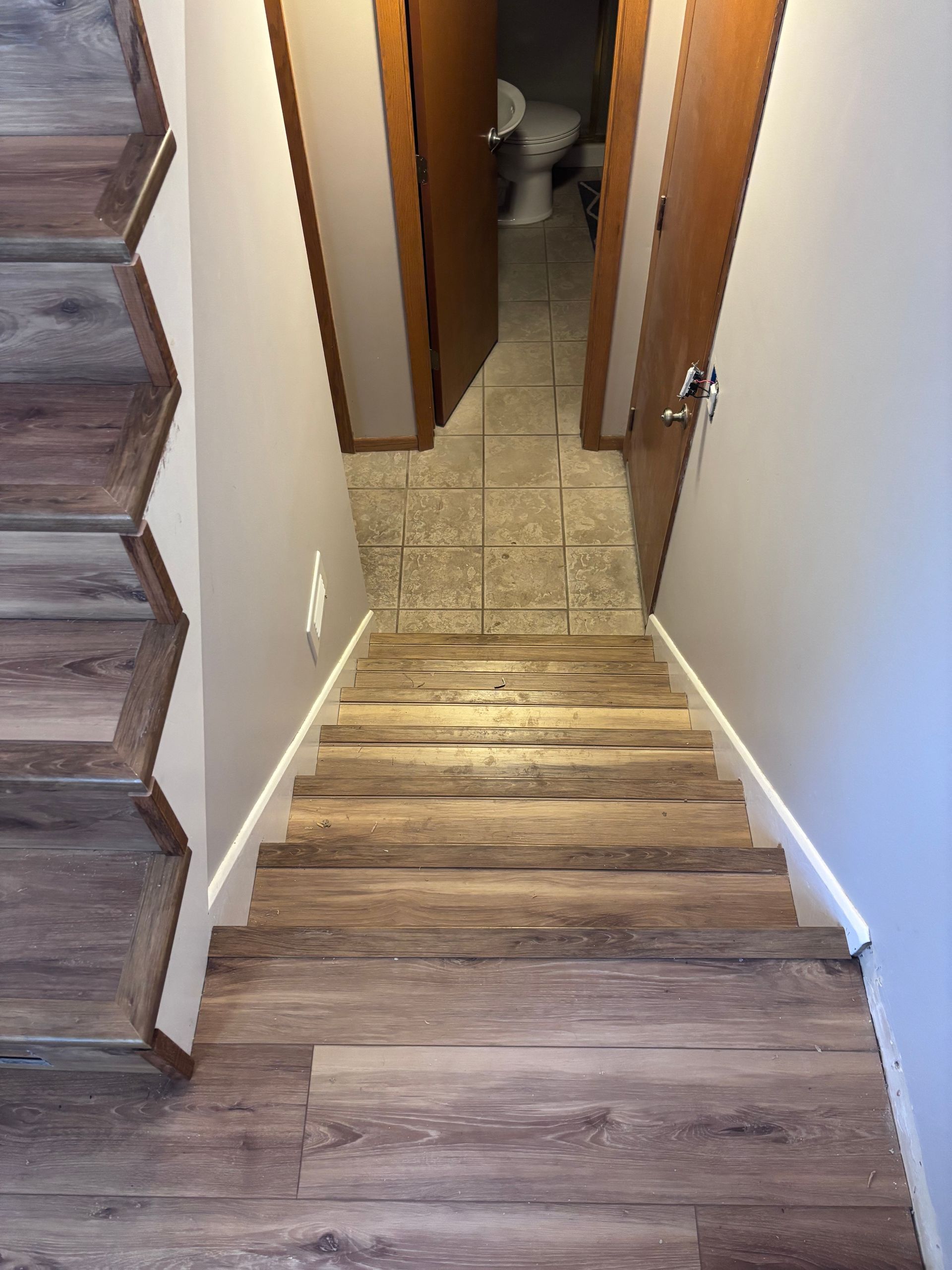 Wooden stairs leading down, with bathroom and door at the end. Beige walls and flooring.
