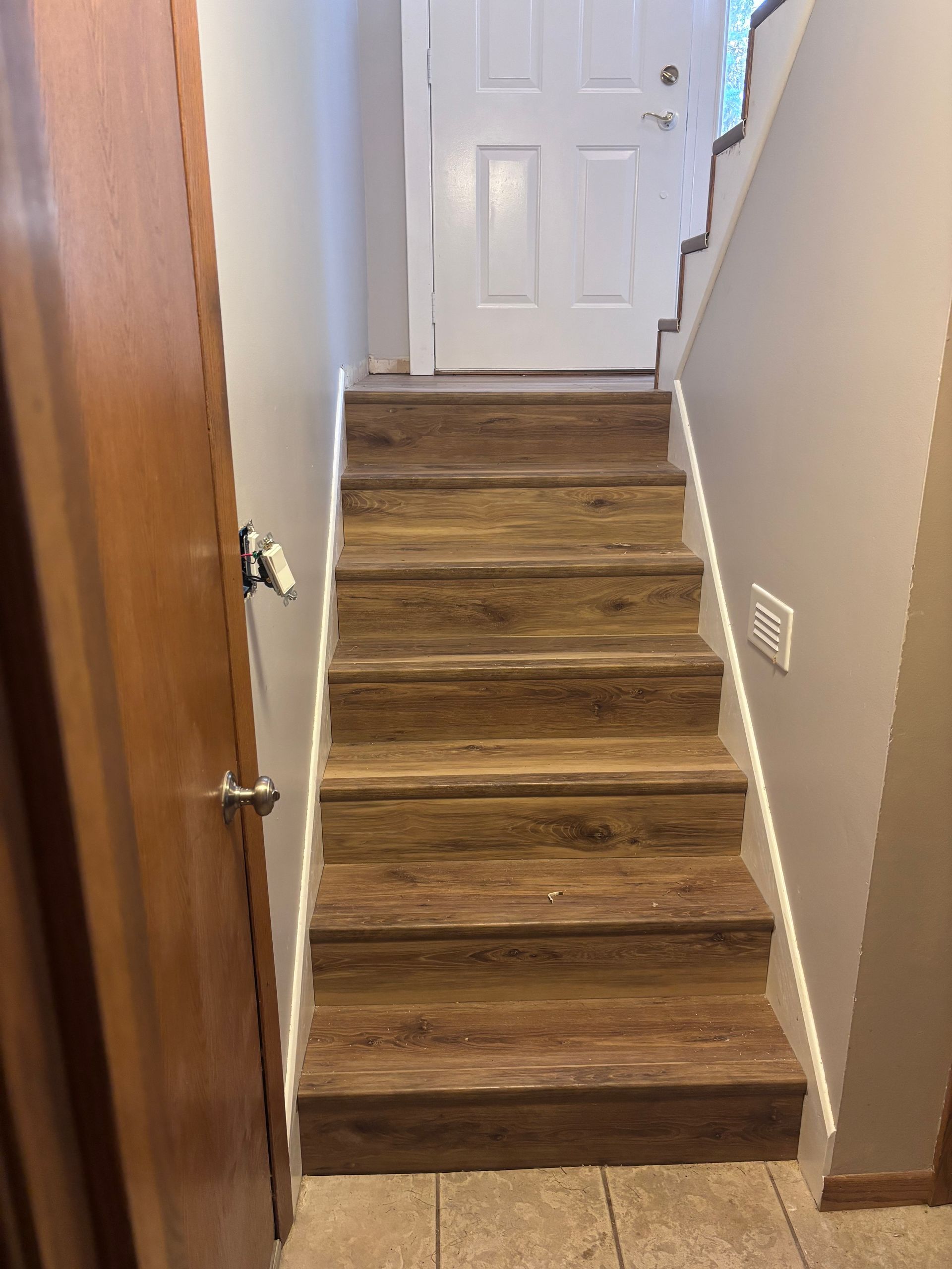 Staircase with wood-look steps leading up to a white door. The walls are neutral colors, with a brown door on the left.