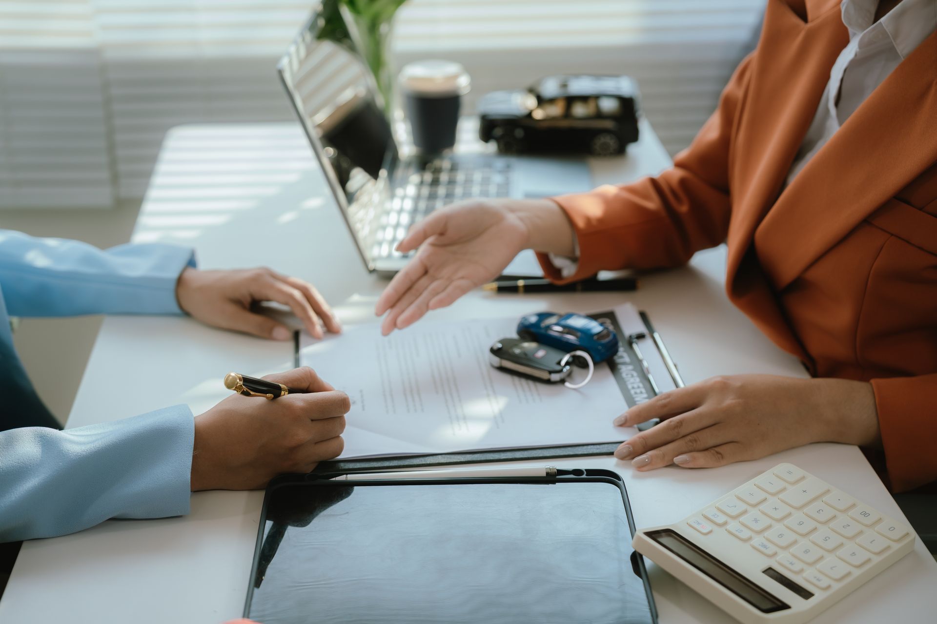 A person signing a contract while another gestures toward the document on a desk with a car key, laptop, and calculator.