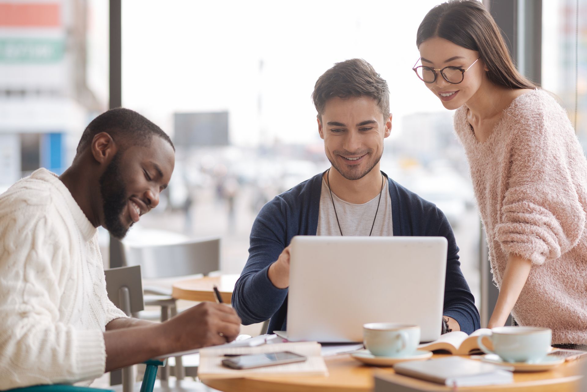 Three people collaborate on a laptop at a sunny cafe, smiling while working together on a project.