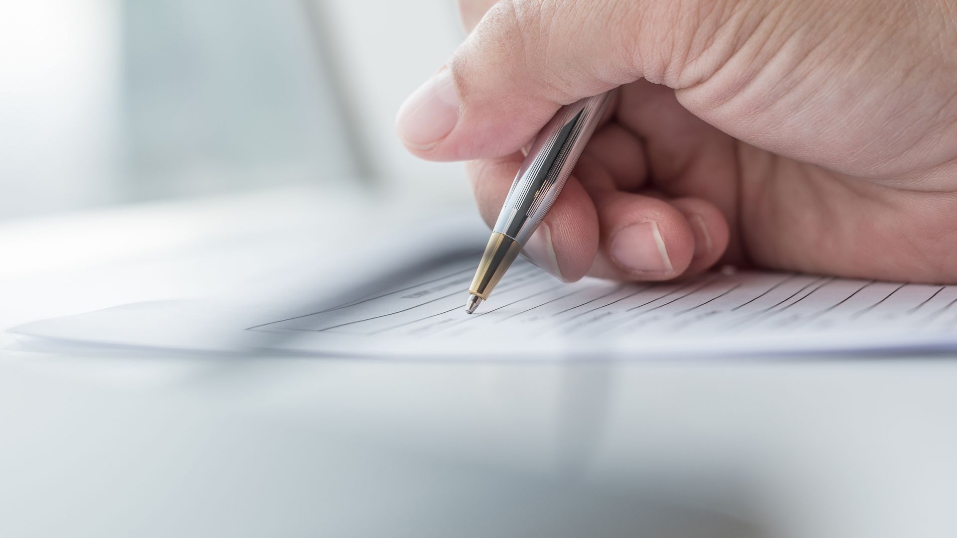 A close-up view of a hand using a metallic pen to fill out a document on a bright, minimalist desk.