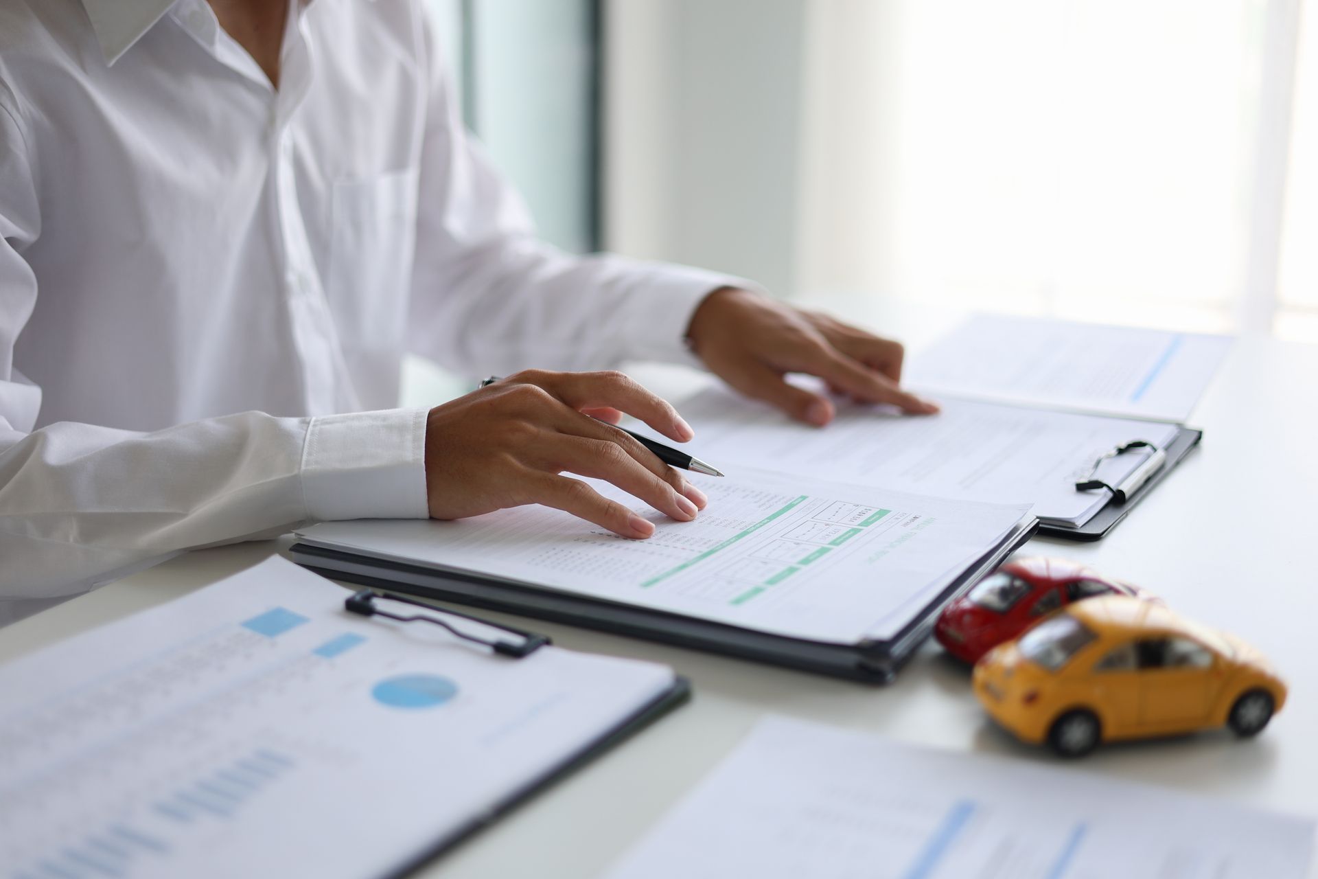 A person in a white shirt sits at a desk with documents, a pen, and two small toy cars, representing car insurance.