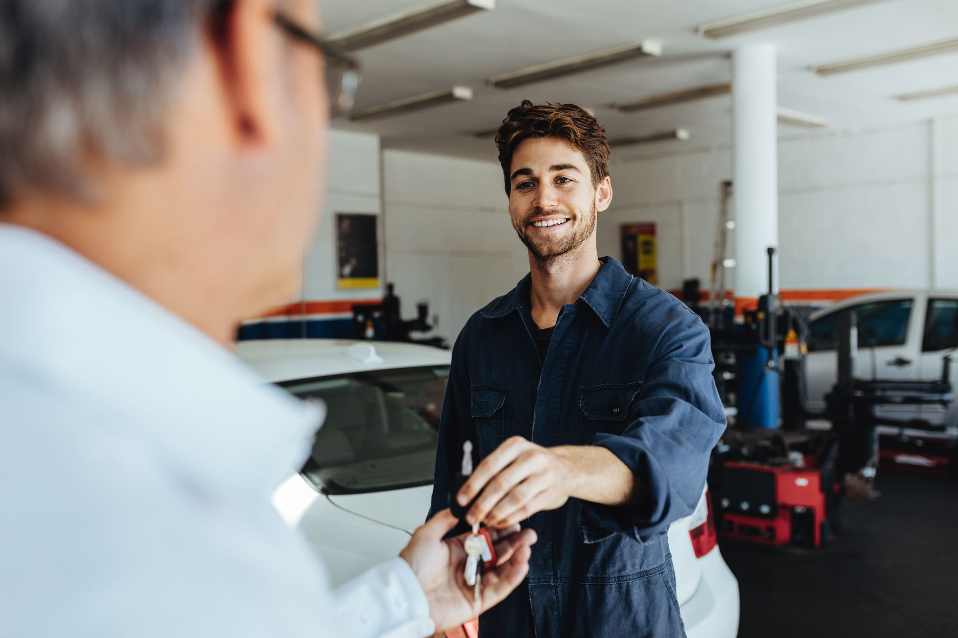 A smiling auto mechanic in a dark blue uniform hands car keys to a customer inside a brightly lit repair shop.