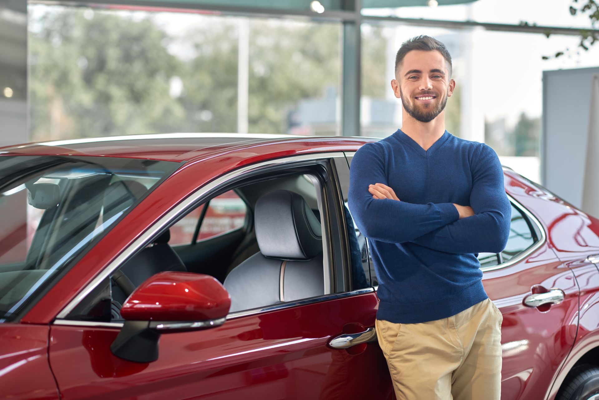 A smiling person with folded arms leans against a red car in a dealership showroom.