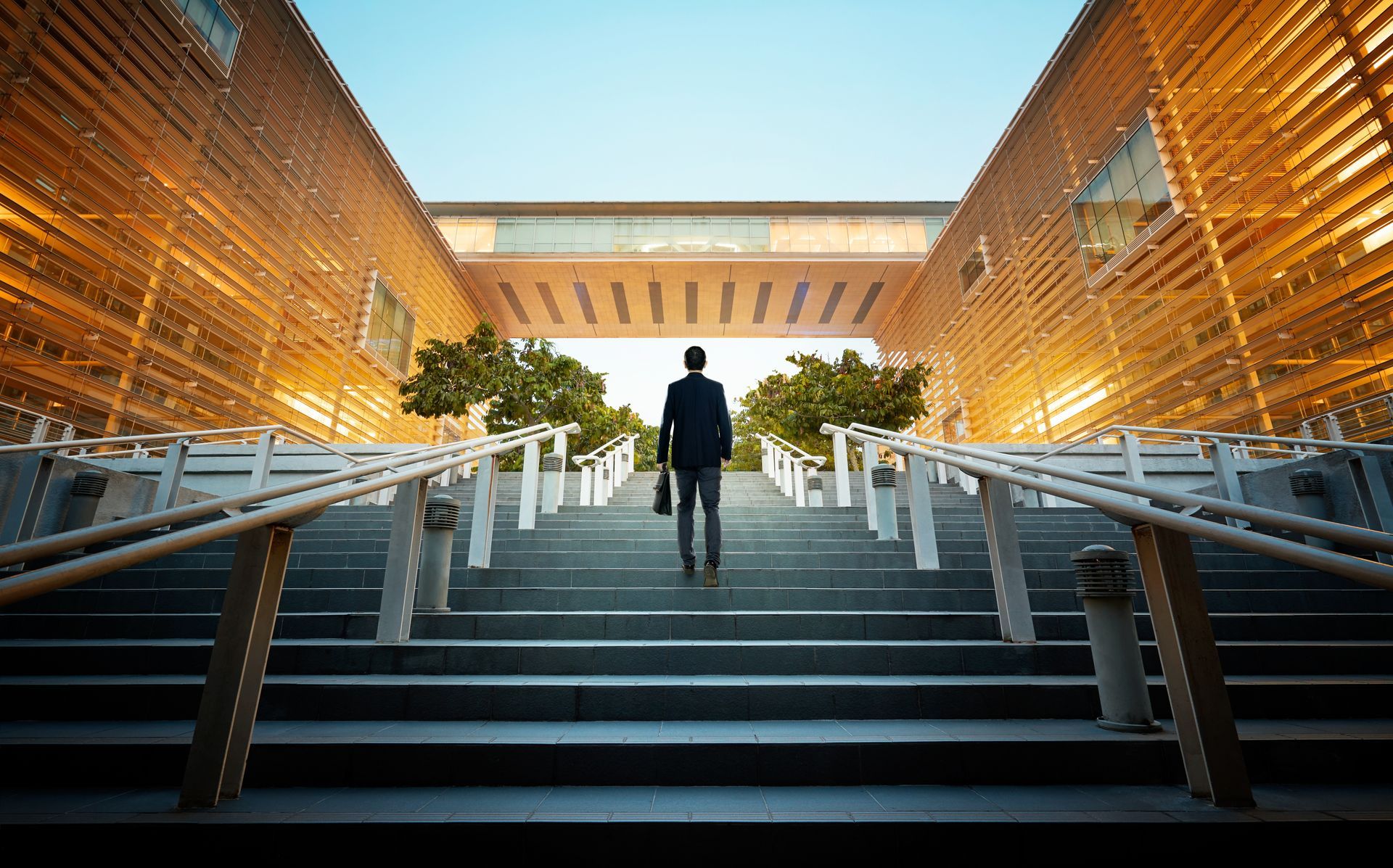A lone figure in a suit walks up wide, stone steps toward a bridge connecting two modern buildings with glowing slats.