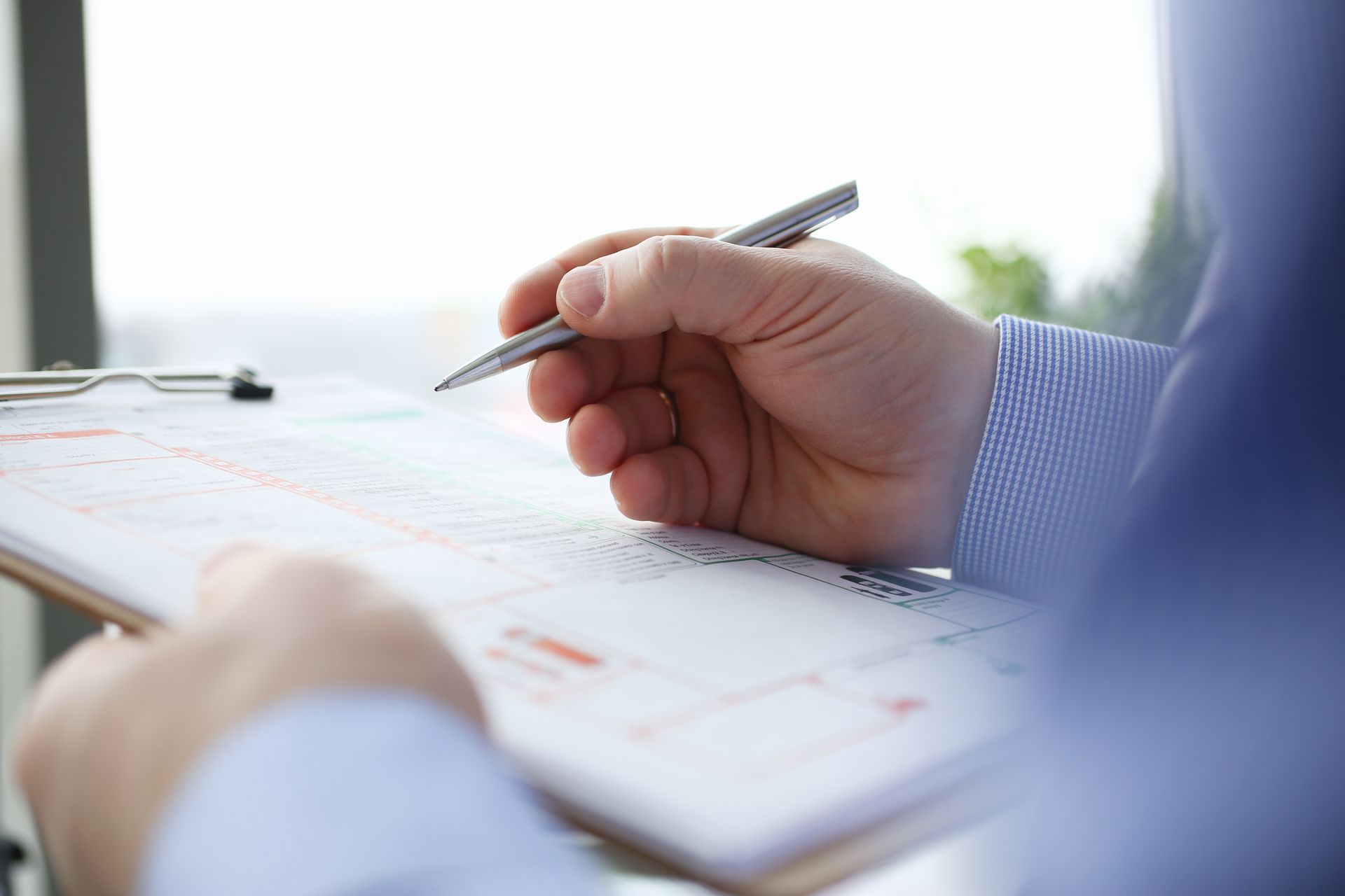 A person in a blue shirt holds a pen while reviewing and marking a document on a clipboard.