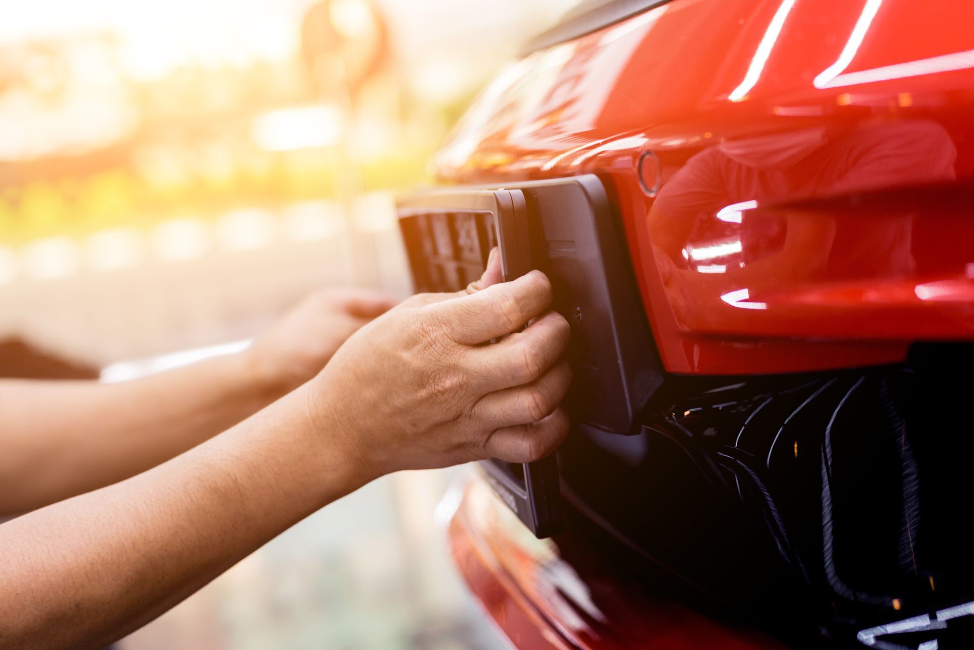 Hands attaching a black license plate frame to the front bumper of a shiny red car in bright daylight.