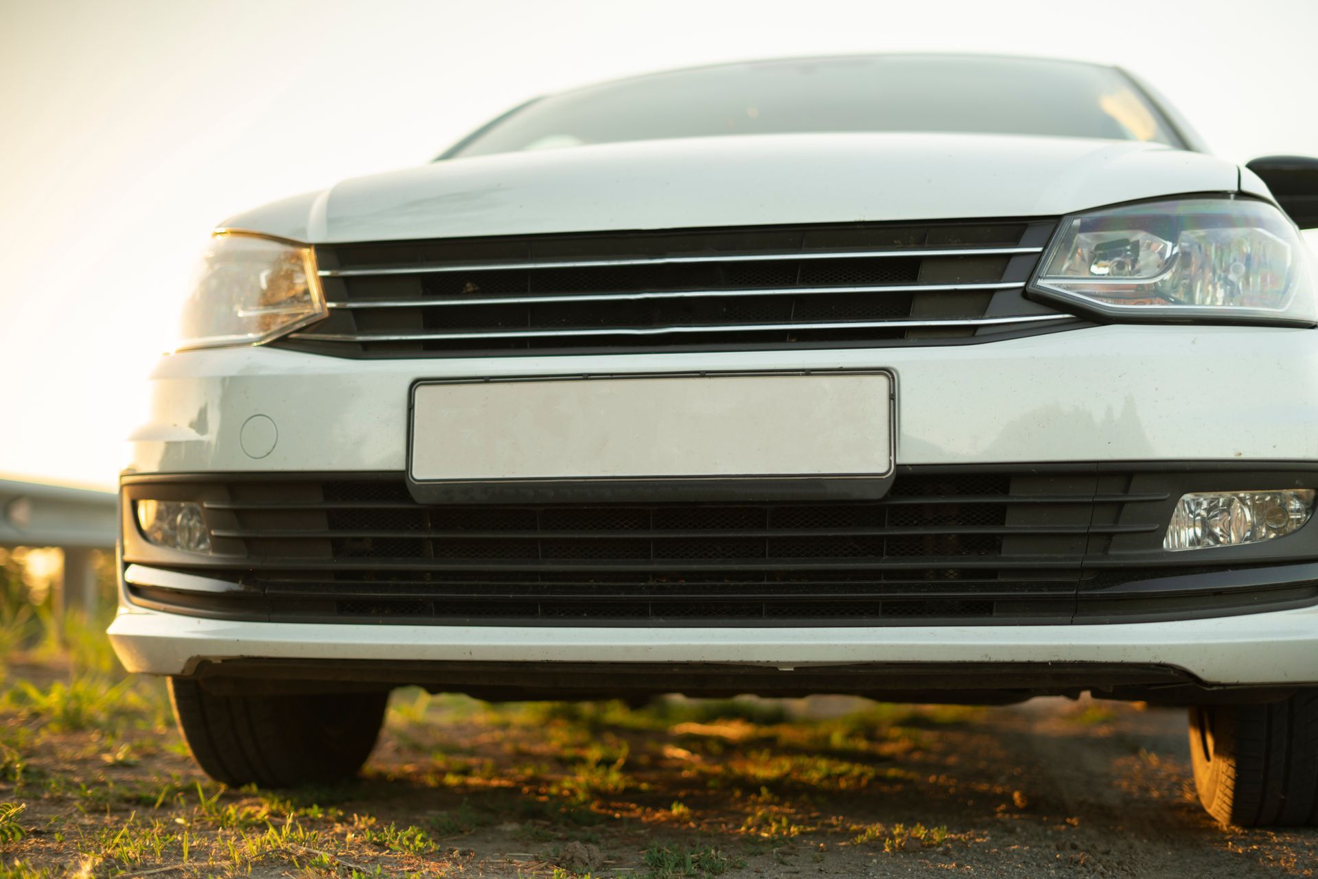 Low-angle view of a white sedan's front grille, headlights, and bumper parked on a grassy dirt area at sunset.