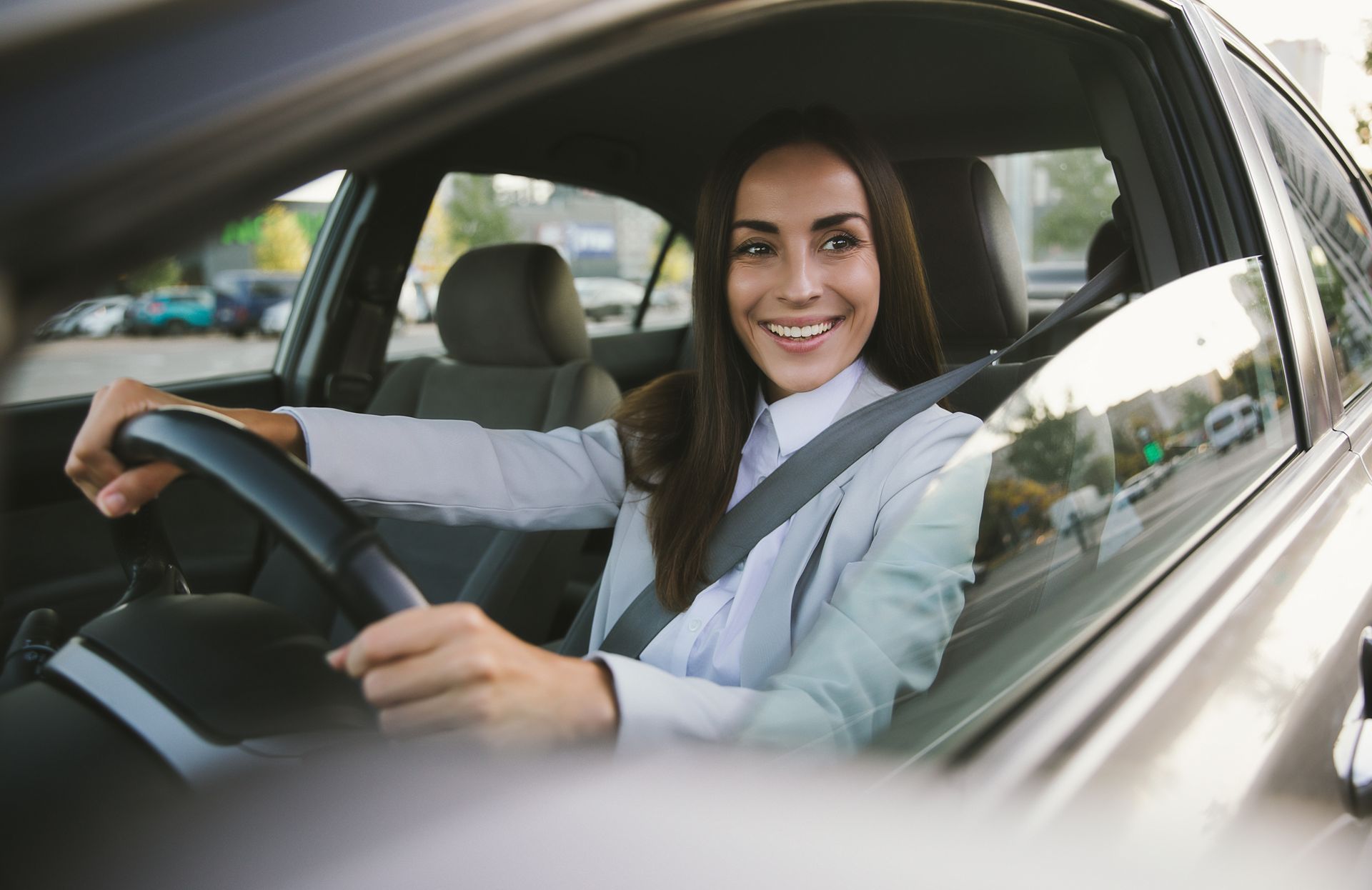 A smiling person in a light-colored shirt driving a car, wearing a seatbelt and holding the steering wheel.
