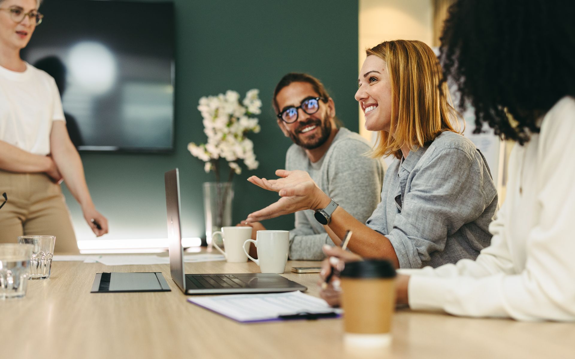 A diverse group of colleagues collaborating in a bright, modern office meeting around a table with a laptop and coffee.