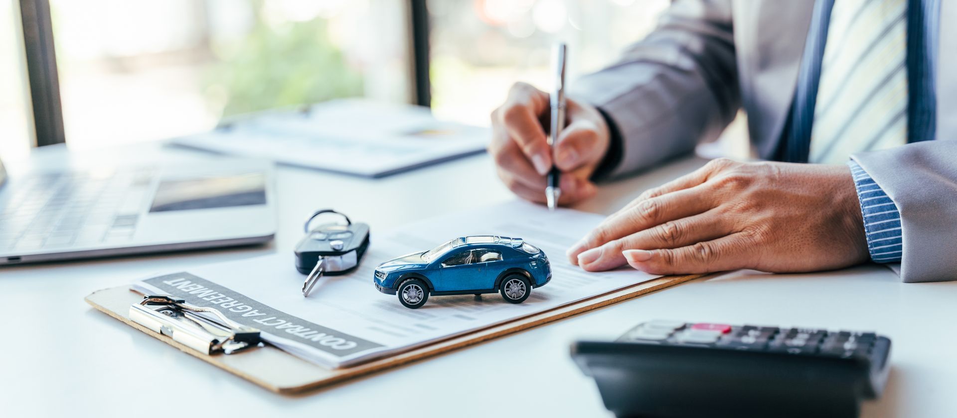 A person in a suit writing on a document at a desk with a toy blue car, car keys, and a calculator.