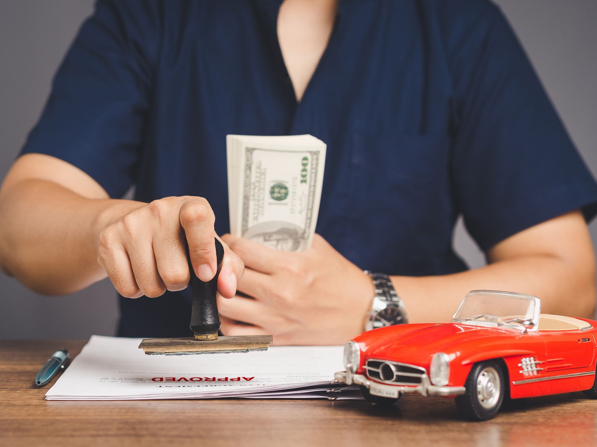 A person stamping a document near a stack of cash and a red toy car on a desk.