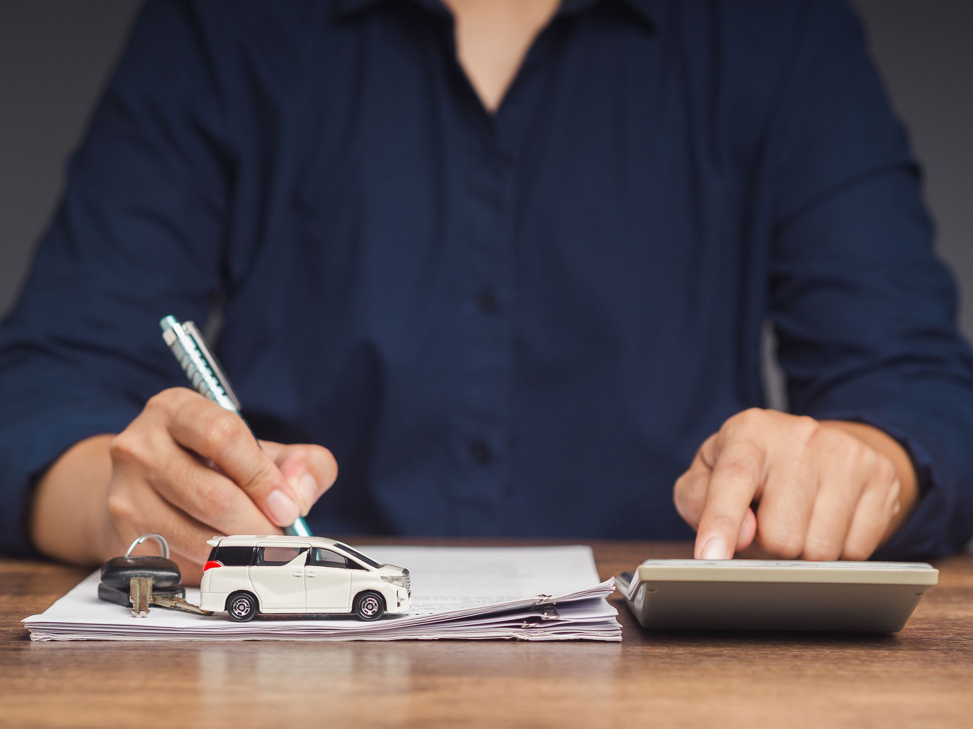 Person writing on paperwork at a desk beside a toy car and calculator Person writing on paperwork at a desk beside a toy car and calculator