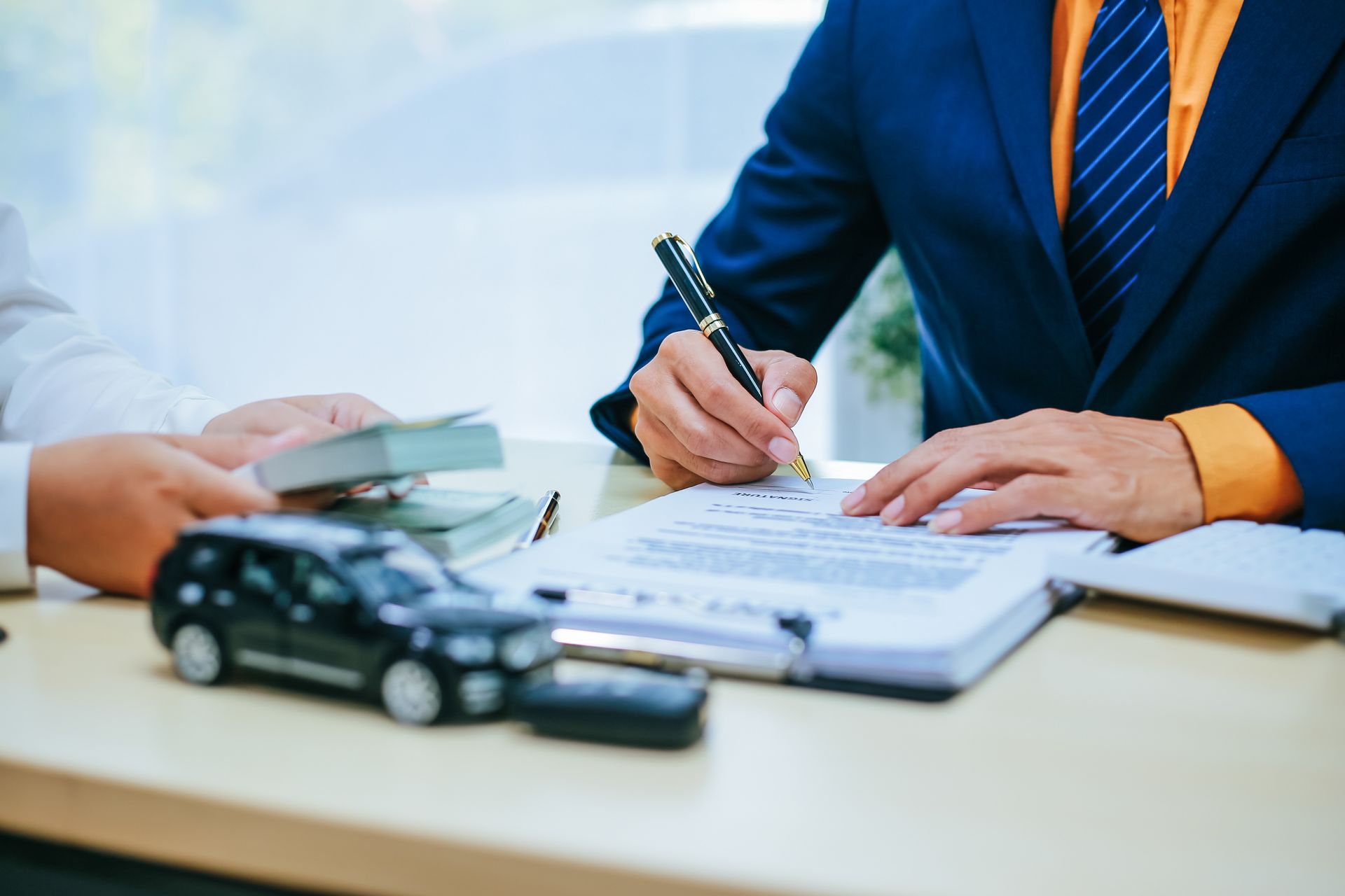 A person in a blue suit signs a document at a desk with a toy car and money present, representing a vehicle purchase.