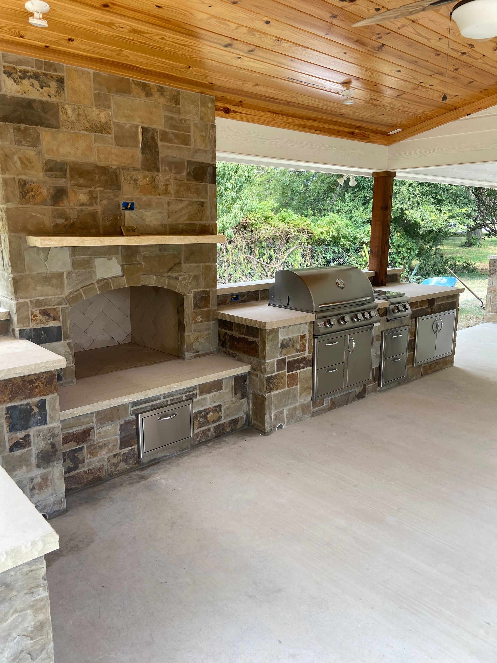 Outdoor kitchen with stone facade, fireplace, stainless steel grill, and beige countertop.