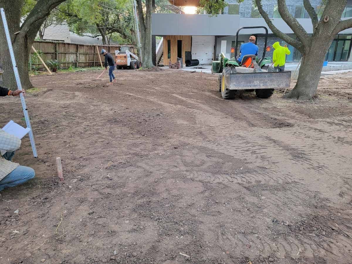Construction site: a roller compacting dirt in a yard, with workers and a building in the background.