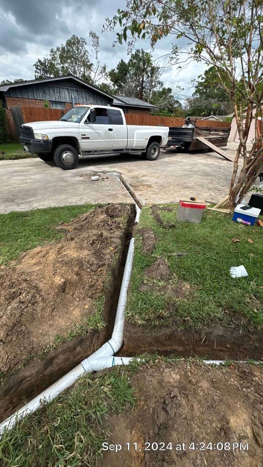 Trench with drainage pipes being installed next to a driveway. A white truck and trailer are parked nearby.