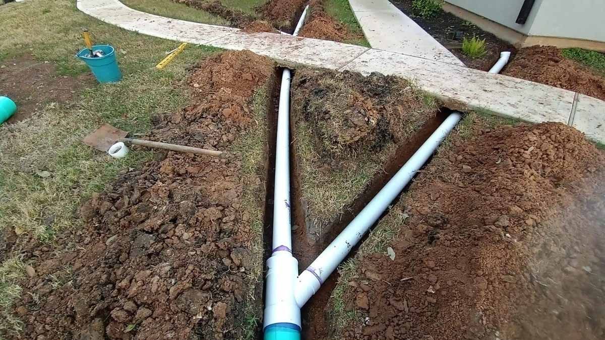 Excavation site with white PVC pipes branching out in a yard, near a walkway.