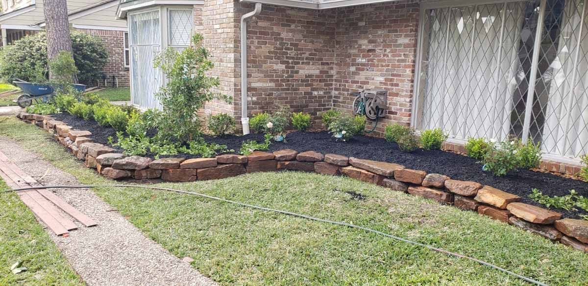 Stone retaining wall borders a flower bed filled with mulch and plants in front of a house.