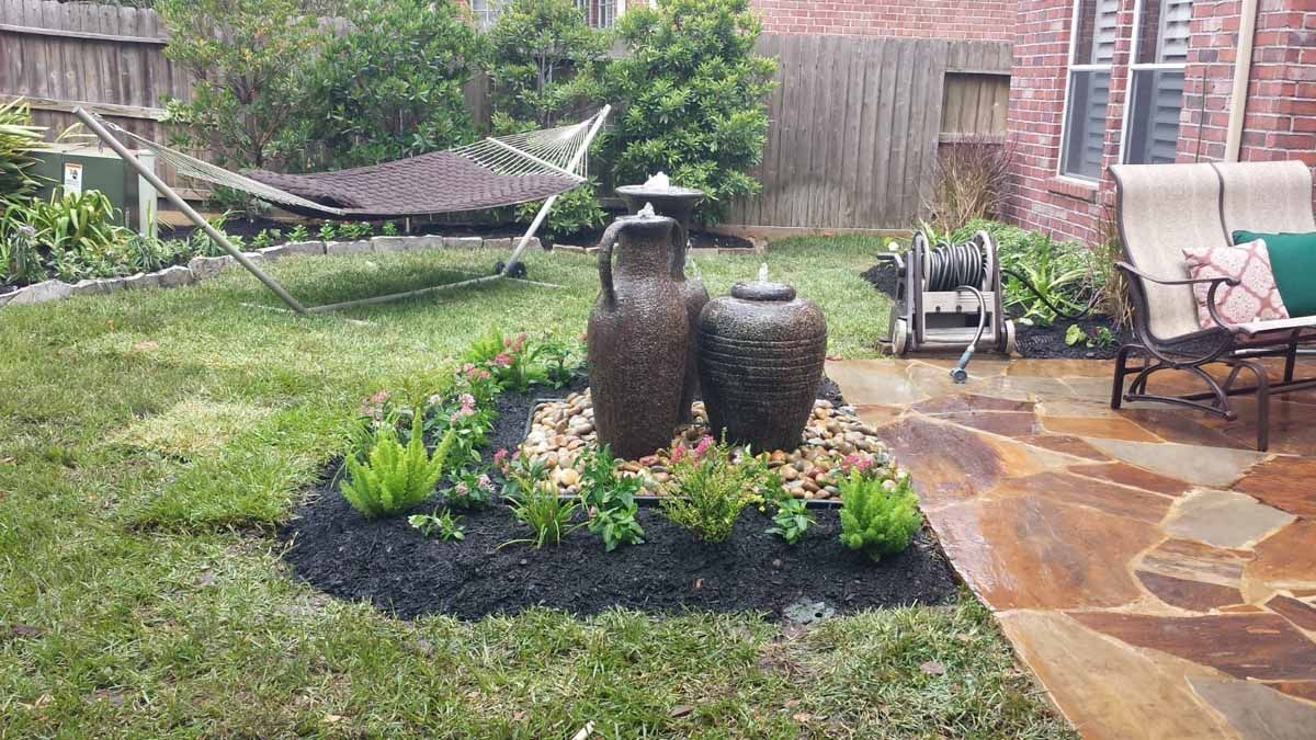Backyard with fountain, hammock, patio furniture, and plants; green grass and brown stone path.