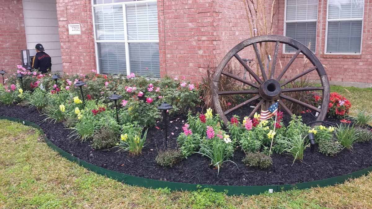 Garden bed with wagon wheel, flowers, and solar lights in front of a brick house.