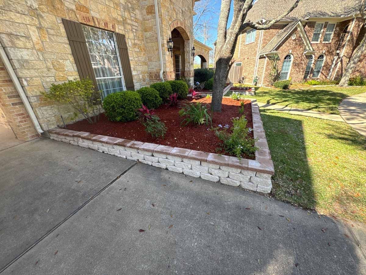Stone planter box with red mulch and plants in front of a house.