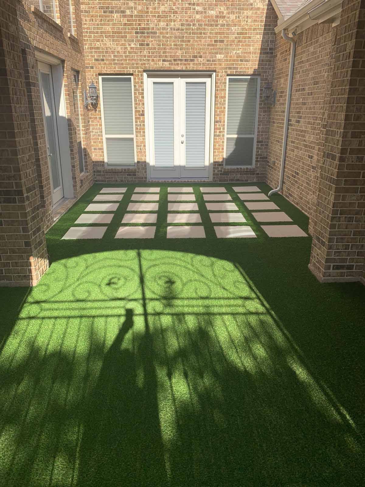 Green artificial turf patio with stone squares leading to double doors under brick walls.